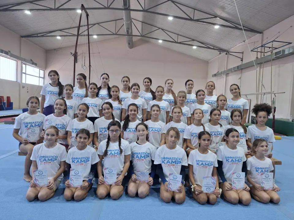 Velika grupa devojčica u majicama gimnastičkog kampa sa sertifikatima u rukama, u gimnastičkoj sali