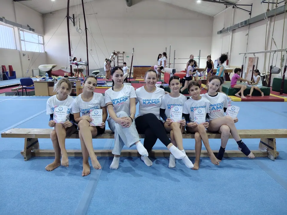 Devojčice i trenerice u majicama gimnastičkog kampa sede na klupi u sali i drže sertifikate